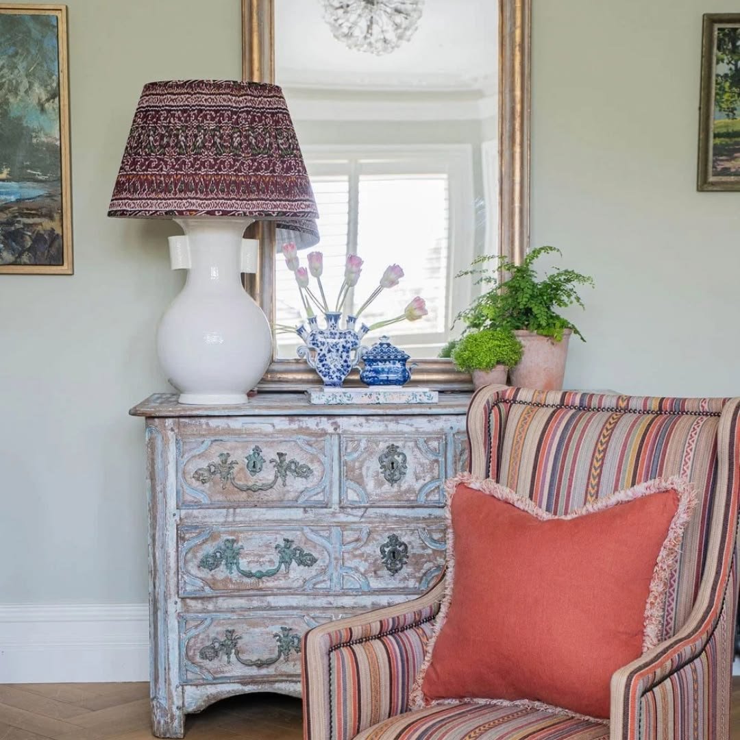 White ceramic lamp with a patterned shade on a painted chest beside a striped armchair