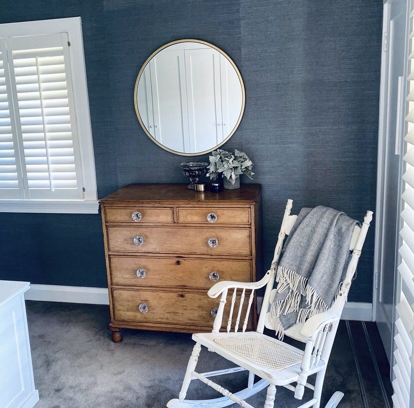 Antique timber chest of drawers with a round mirror and a white rocking chair against blue grasscloth, The Style Workshop