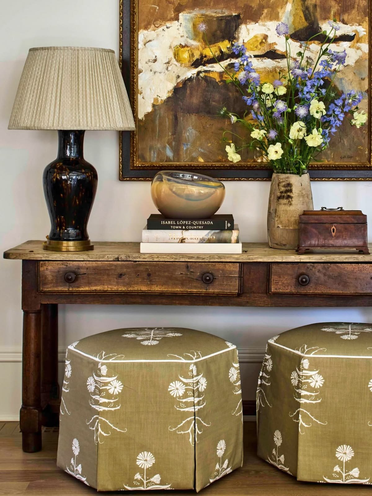 Timber console with artwork, a table lamp, books and a pair of upholstered ottomans